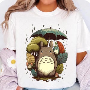 Op de afbeelding: Wit T-shirt met een cartoon illustratie van een groot, grijs wezen met een cr&egrave;mekleurige buik, die een groene paraplu vasthoudt. Het ontwerp bevat bomen, bladeren en regendruppels.