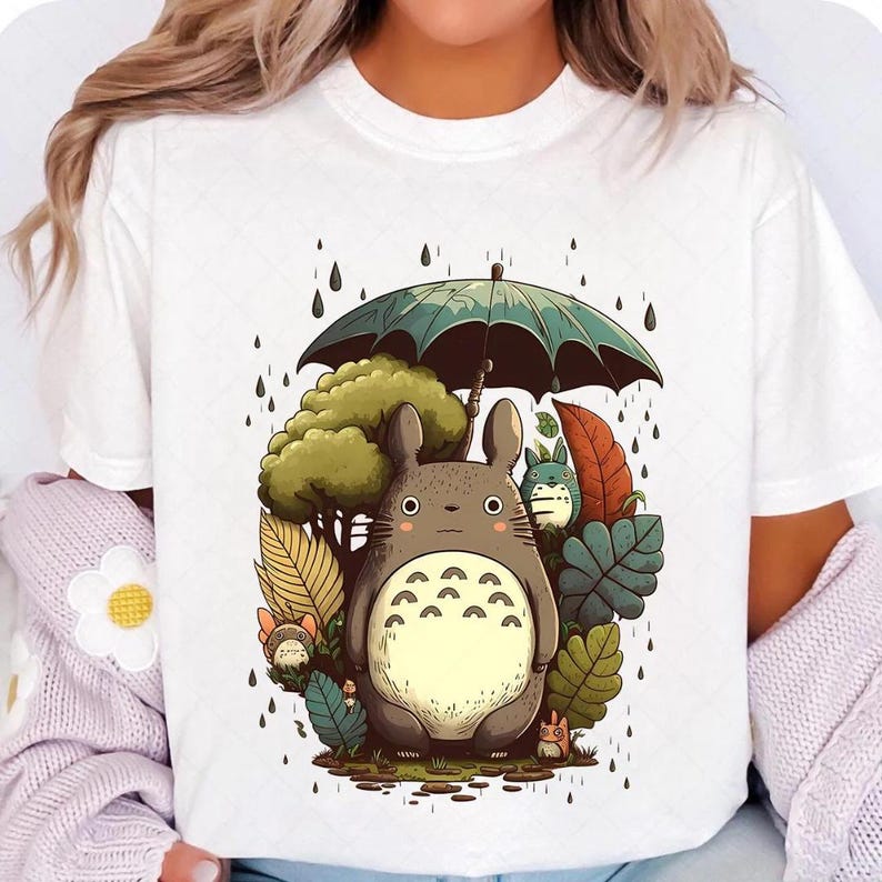 Op de afbeelding: Wit T-shirt met een cartoon illustratie van een groot, grijs wezen met een cr&egrave;mekleurige buik, die een groene paraplu vasthoudt. Het ontwerp bevat bomen, bladeren en regendruppels.