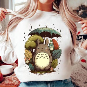 Op de afbeelding: Witte sweatshirt met een cartoon illustratie van Totoro onder een paraplu in de regen. Het ontwerp bevat een groot grijs Totoro-personage, groene bomen en kleurrijke bladeren. Regendruppels vallen rond de personages.