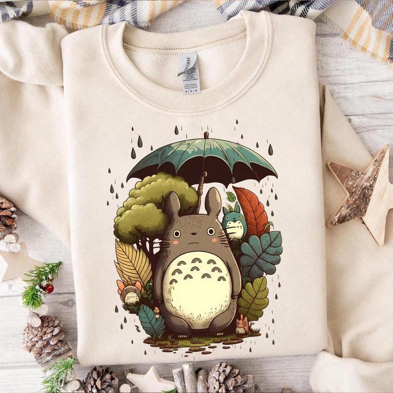 Op de afbeelding: Cr&egrave;mekleurige sweatshirt met een kleurrijke illustratie van Totoro onder een paraplu in de regen. Het ontwerp bevat een groene boom, diverse bladeren en andere personages uit de animatiefilm. Een gezellig, casual kledingstuk.