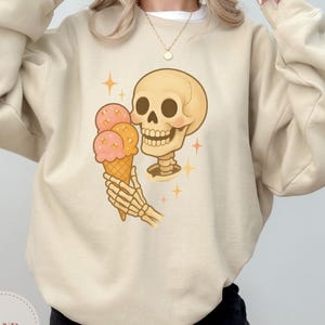 Op de afbeelding: Zandkleurige sweatshirt met een grillige illustratie van een skelet dat een ijshoorntje vasthoudt met roze en oranje bollen. Het ontwerp bevat steraccenten en een lachende schedel. Het woord "SAND" staat linksonder.