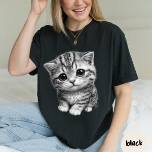Peut inclure: T-shirt noir avec une illustration d&eacute;taill&eacute;e d'un chaton gris et blanc aux grands yeux expressifs. Le chaton est le point central, rendu dans un style r&eacute;aliste. Le mot "black" est dans un cercle en bas &agrave; droite.