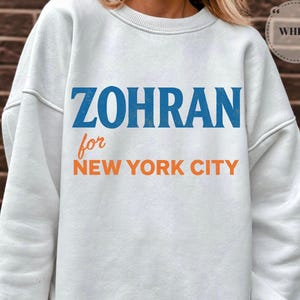 May include: White sweatshirt with the text "ZOHRAN for NEW YORK CITY" in blue and orange lettering. The sweatshirt has a crew neck and long sleeves. The word "WHITE" is in a white circle in the upper right corner.