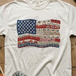 Pode incluir: Camiseta de cor marfim com um design bordado da bandeira americana. A bandeira incorpora estrelas, listras e símbolos patrióticos. A camiseta é feita de um material macio. A palavra "Ivory" está impressa no canto superior direito.