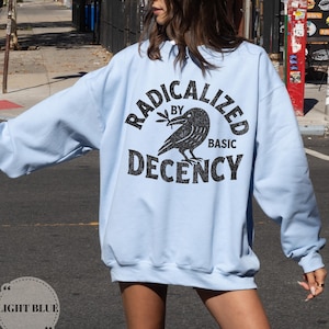 Pode incluir: Camisola azul clara com o texto preto "RADICALIZED BY BASIC DECENCY". Um gráfico de corvo preto está centrado abaixo do texto. A camisola é azul clara e tem mangas compridas.