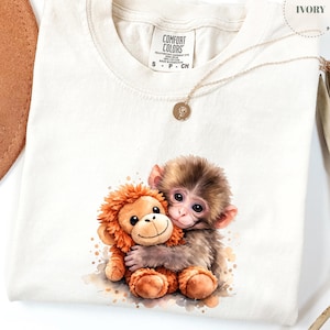 May include: Ivory-colored t-shirt featuring a watercolor illustration of two hugging monkeys. One is a plush toy, the other a real monkey. A gold necklace with a small pendant rests on the shirt. The shirt has a Comfort Colors tag.