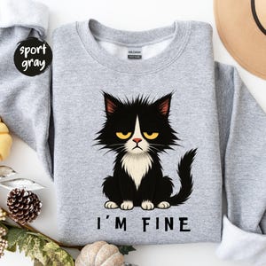 May include: A light grey sweatshirt featuring a black and white cat illustration with a grumpy expression and yellow eyes. The text "I'M FINE" is printed below the cat. The sweatshirt is labelled "sport gray".