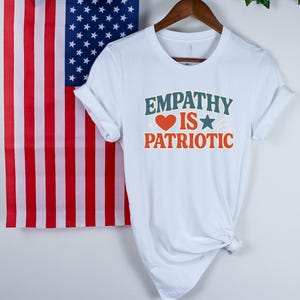 May include: White t-shirt with the text "EMPATHY IS PATRIOTIC" in teal and orange. The design includes a red heart and a blue star. An American flag hangs in the background.