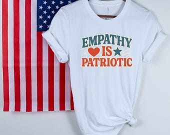 May include: White t-shirt with the text "EMPATHY IS PATRIOTIC" in teal and orange. The design includes a red heart and a blue star. An American flag hangs in the background.