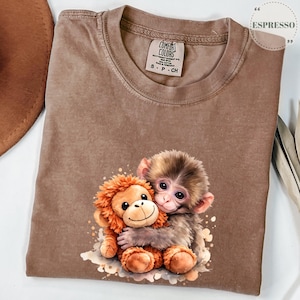 Może przedstawiać: Koszulka Comfort Colors w kolorze espresso z grafiką przedstawiającą małą małpkę obejmującą pluszową małpkę. Koszulka ma okrągły dekolt i kr&oacute;tkie rękawy. Koszulka ma małą metkę z napisem "Comfort Colors".