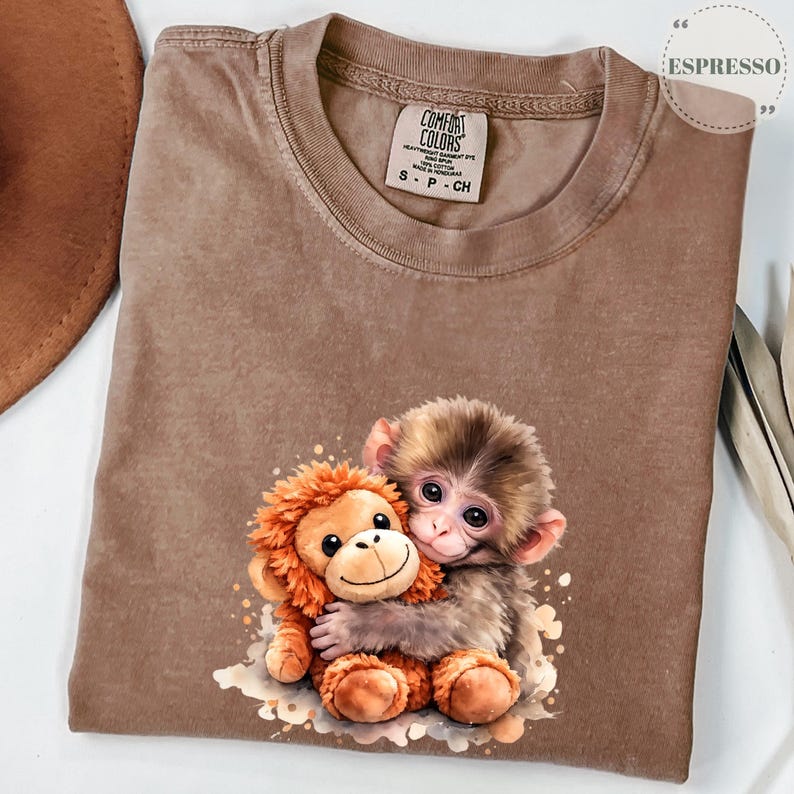 Może przedstawiać: Koszulka Comfort Colors w kolorze espresso z grafiką przedstawiającą małą małpkę obejmującą pluszową małpkę. Koszulka ma okrągły dekolt i kr&oacute;tkie rękawy. Koszulka ma małą metkę z napisem "Comfort Colors".