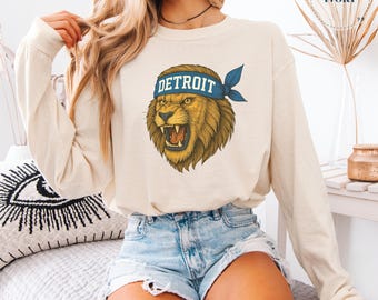 Op de afbeelding: Ivoor lange mouwen sweatshirt met een brullende leeuw grafische afbeelding met een blauwe hoofdband met de tekst "DETROIT". De leeuw is in bruine en gouden tinten. Het sweatshirt wordt gedragen met een spijkerbroek short.