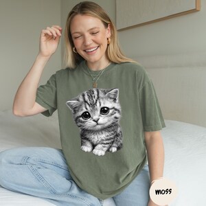 Peut inclure: T-shirt vert olive avec un grand motif d'un chaton gris et blanc aux grands yeux. Le t-shirt a une coupe d&eacute;contract&eacute;e et est port&eacute; avec un jean bleu. Un petit autocollant rond avec le mot "moss" est visible.