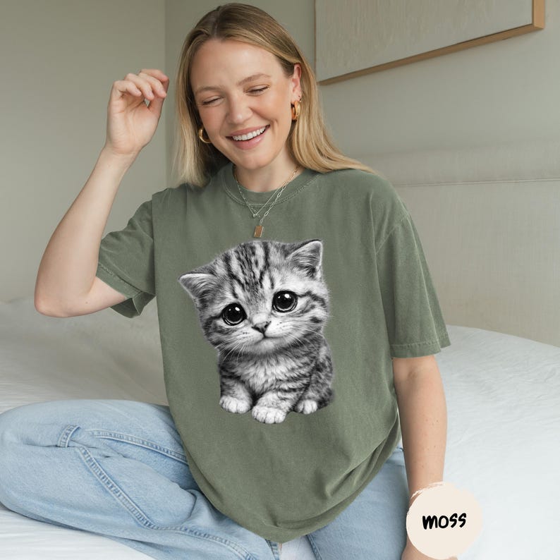 Peut inclure: T-shirt vert olive avec un grand motif d'un chaton gris et blanc aux grands yeux. Le t-shirt a une coupe d&eacute;contract&eacute;e et est port&eacute; avec un jean bleu. Un petit autocollant rond avec le mot "moss" est visible.