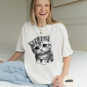 Peut inclure: T-shirt blanc avec une illustration en noir et blanc d'un chaton aux grands yeux. Le mot "white" est imprim&eacute; en bas &agrave; droite du t-shirt. Le t-shirt est de style d&eacute;contract&eacute;.