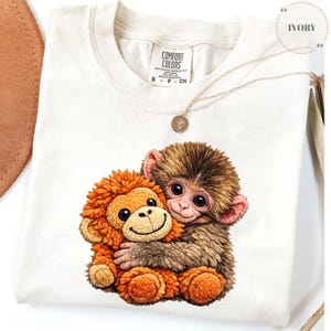 Puede incluir: Camiseta de color marfil con un gráfico de dos monos de dibujos animados abrazándose. Los monos tienen pelaje marrón y naranja y ojos grandes. Un collar de oro con un pequeño colgante descansa sobre la camiseta. La camiseta está etiquetada como "Comfort Colors".