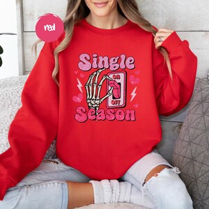 Puede incluir: Sudadera roja con la frase "Single Season" en letras de purpurina rosa. Se muestra una mano de esqueleto accionando un interruptor de "encendido/apagado". El dise&ntilde;o tambi&eacute;n incluye corazones rosas y rayos.
