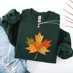 Op de afbeelding: Donkergroene sweatshirt met een groot, kleurrijk esdoornblad-ontwerp. Het blad bestaat uit verschillende patchworkpatronen in tinten oranje, geel en groen. Een zilveren ketting met een kleine hanger ligt op de sweatshirt.