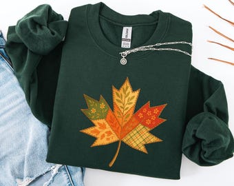Puede incluir: Sudadera verde oscuro con un diseño de hoja de arce grande y colorida. La hoja está compuesta por varios parches estampados en tonos naranja, amarillo y verde. Un collar plateado con un pequeño colgante está sobre la sudadera.