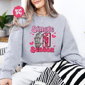 Puede incluir: Una sudadera gris claro con las palabras "Single Season" en rosa, con una mano de esqueleto accionando un interruptor. Corazones rosas y rayos acent&uacute;an el dise&ntilde;o. La sudadera tiene cuello redondo.