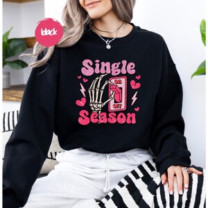 Puede incluir: Sudadera negra con la frase "Single Season" en rosa, con una mano de esqueleto accionando un interruptor. Corazones rosas y rayos acent&uacute;an el dise&ntilde;o. Un c&iacute;rculo rosa con la palabra "black" est&aacute; en la parte superior izquierda.