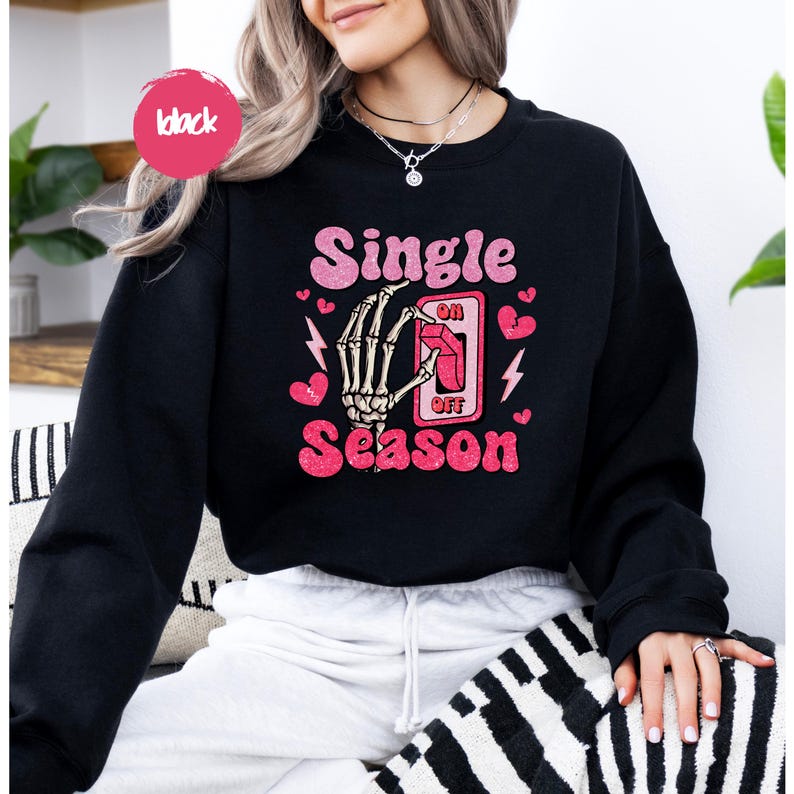 Puede incluir: Sudadera negra con la frase "Single Season" en rosa, con una mano de esqueleto accionando un interruptor. Corazones rosas y rayos acent&uacute;an el dise&ntilde;o. Un c&iacute;rculo rosa con la palabra "black" est&aacute; en la parte superior izquierda.
