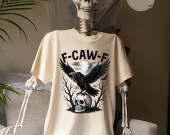Könnte beinhalten: Elfenbeinfarbenes T-Shirt mit einem schwarzen Raben, der auf einem Totenkopf sitzt, mit dem Text "F-CAW-F" darüber. Das Design umfasst einen Vollmond und kahle Äste, die ein gruseliges Halloween-Thema erzeugen.