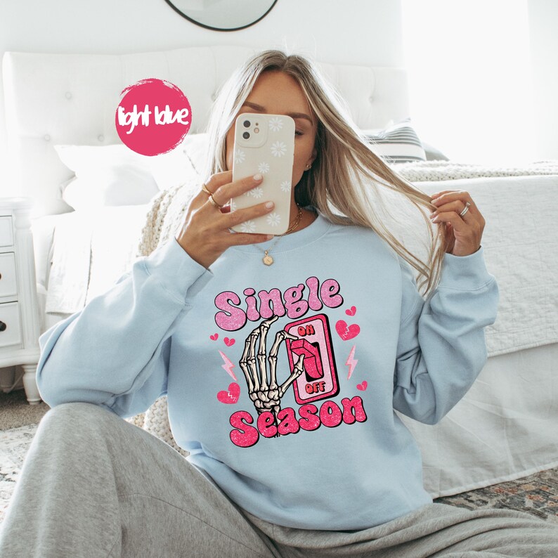 Puede incluir: Sudadera azul claro con el texto "Single Season" en rosa, con una mano de esqueleto accionando un interruptor. Corazones rosas y rayos acent&uacute;an el dise&ntilde;o. La sudadera es de color azul claro liso.