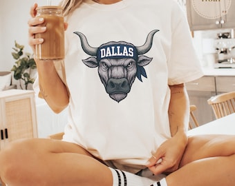 Op de afbeelding: Ivoor kleurig T-shirt met een gedetailleerde illustratie van een stierenkop met het woord "DALLAS" op een blauwe hoofdband. De stier heeft grote hoorns en een serieuze uitdrukking. Het shirt heeft een relaxte pasvorm.