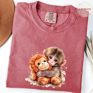 Może przedstawiać: Koszulka Comfort Colors w kolorze przydymionego r&oacute;żu z akwarelową ilustracją dw&oacute;ch małp obejmujących się. Jedna to pluszowa zabawka, druga prawdziwa małpa. Koszulka ma okrągły dekolt i małą metkę z napisem "S P CH".