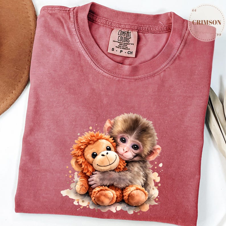 Może przedstawiać: Koszulka Comfort Colors w kolorze przydymionego r&oacute;żu z akwarelową ilustracją dw&oacute;ch małp obejmujących się. Jedna to pluszowa zabawka, druga prawdziwa małpa. Koszulka ma okrągły dekolt i małą metkę z napisem "S P CH".