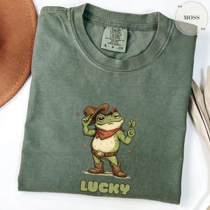 Puede incluir: Camiseta verde salvia con una rana de dibujos animados con sombrero de vaquero, pañuelo rojo y botas, sosteniendo un trébol de cuatro hojas. La palabra "LUCKY" está impresa en una fuente marrón en la parte inferior. La camiseta tiene una etiqueta Comfort Colors.
