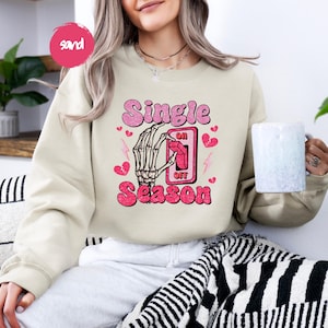 Puede incluir: Una sudadera color arena con las palabras "Single Season" en rosa, con una mano de esqueleto accionando un interruptor de "encendido/apagado". Corazones rosas y rayos acent&uacute;an el dise&ntilde;o. La sudadera es de estilo casual y c&oacute;modo.