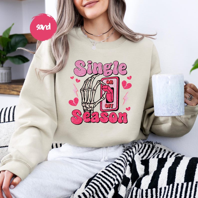 Puede incluir: Una sudadera color arena con las palabras "Single Season" en rosa, con una mano de esqueleto accionando un interruptor de "encendido/apagado". Corazones rosas y rayos acent&uacute;an el dise&ntilde;o. La sudadera es de estilo casual y c&oacute;modo.