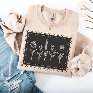 May include: A sand-colored sweatshirt with a black rectangular patch featuring white line drawings of flowers. The patch is outlined with a stitched border. A delicate necklace with a small pendant rests on the sweatshirt. The sweatshirt is folded on top of a pair of blue jeans.