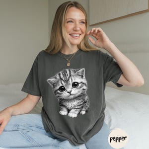 Peut inclure: T-shirt gris fonc&eacute; avec une illustration en noir et blanc d'un chaton aux grands yeux. Le t-shirt a des manches courtes et une coupe d&eacute;contract&eacute;e. Le mot "pepper" est imprim&eacute; en bas &agrave; droite.