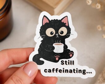 Può includere: Un adesivo fustellato raffigurante un gatto nero soffice con grandi occhi e orecchie rosa, seduto mentre tiene una piccola tazza bianca da cui esce vapore. Il testo sottostante recita Still caffeinating in un carattere nero, grassetto e sans-serif.