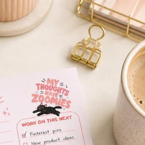 May include: A desk scene with a planner featuring the text "My Thoughts Have Zoomies" and a black cat illustration. A gold binder clip, a cup of coffee, and a ceramic container are also visible. The planner includes a checklist with "Pinterest pins" checked.