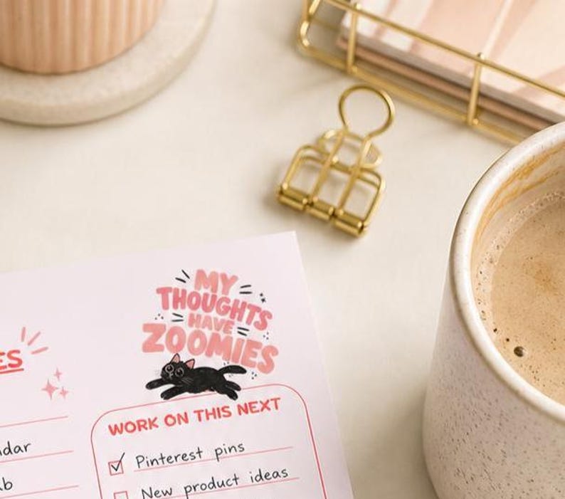 May include: A desk scene with a planner featuring the text "My Thoughts Have Zoomies" and a black cat illustration. A gold binder clip, a cup of coffee, and a ceramic container are also visible. The planner includes a checklist with "Pinterest pins" checked.