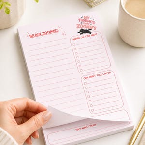 May include: A pink notepad with the text "BRAIN ZOOMIES" and "MY THOUGHTS HAVE ZOOMIES". The notepad has sections for "WORK ON THIS NEXT" and "CAN WAIT TILL LATER". A hand is shown lifting a page. A gold pen and a cup of coffee are in the background.