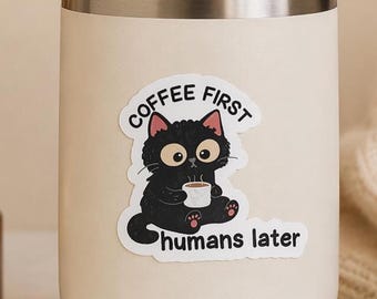 May include: A cream-colored stainless steel tumbler with a clear lid featuring a sticker of a black cat holding a white coffee mug. The text above the cat reads COFFEE FIRST and the text below the cat reads humans later in black lettering.