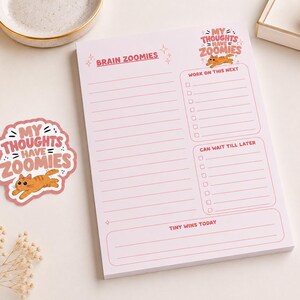 May include: A light pink notepad featuring the text BRAIN ZOOMIES at the top, a section for WORK ON THIS NEXT, a section for CAN WAIT TILL LATER, and a section for TINY WINS TODAY. A matching sticker shows an orange cat with the text MY THOUGHTS HAVE ZOOMIES.