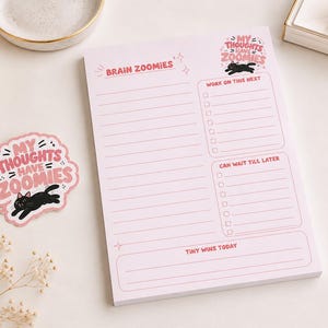 May include: A light pink notepad and a matching sticker featuring a black cat illustration and the text "MY THOUGHTS HAVE ZOOMIES". The notepad includes sections labelled "BRAIN ZOOMIES", "WORK ON THIS NEXT", "CAN WAIT TILL LATER", and "TINY WINS TODAY".