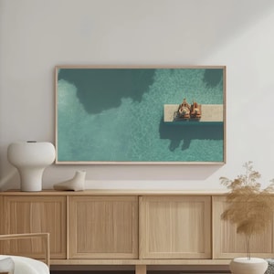 Op de afbeelding: Een ingelijste print toont een luchtfoto van een turquoise zwembad met een kleine steiger en twee figuren. Het kunstwerk wordt weergegeven boven een licht houten kast, met een witte vaas en decoratieve elementen.