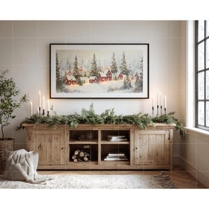 May include: A rustic wooden cabinet with a framed winter landscape painting above it. The painting depicts red houses in a snowy forest. Lit candles and a green garland decorate the cabinet, creating a cozy, festive atmosphere.