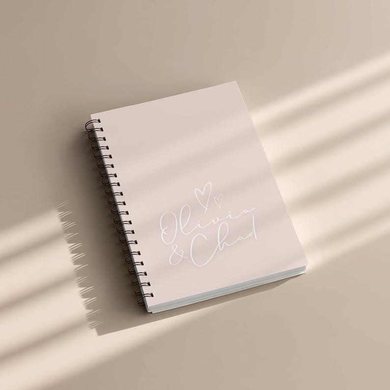 May include: A spiral-bound notebook with a light pink cover. The cover features the names "Olivia & Chad" in a delicate script font, along with two small hearts. The notebook is lying on a neutral surface, with shadows from sunlight.