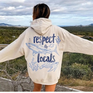 May include: Cream-colored hoodie with a blue graphic of a shark, fish, and turtle, along with the text "I respect the locals." The hoodie has a hood and long sleeves. The background shows a scenic view.