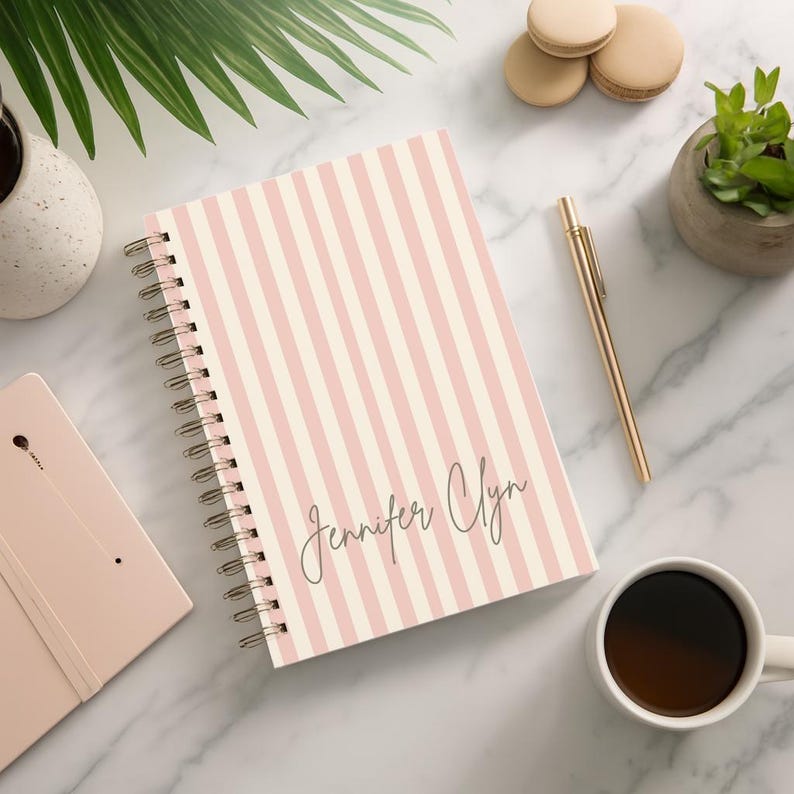 May include: A spiral-bound notebook with a pink and white striped cover. The notebook has the name "Jennifer Clyn" in cursive script. A gold pen, a cup of coffee, and a small potted plant are also visible.