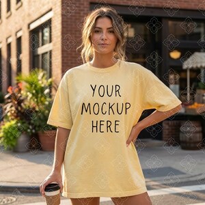 May include: A pale yellow t-shirt with the text "YOUR MOCKUP HERE" in black. The model is holding a coffee cup. The t-shirt is a loose fit and has short sleeves. The background includes a brick building and greenery.