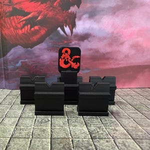 May include: A set of black plastic tabletop gaming card holders with V-shaped notches. One holder features a raised red dragon ampersand logo. These holders are designed to stand upright and display game cards during tabletop role-playing sessions.
